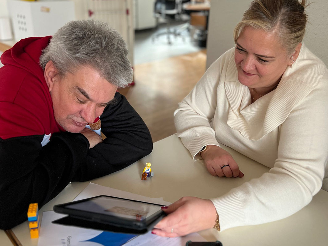 Eine Frau zeigt einem Mann etwas auf einem Tablet. Sie sitzen zusammen am Tisch. 